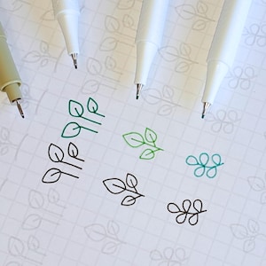 Puede incluir: Se muestran cuatro marcadores blancos de punta fina y un marcador beige con tinta negra sobre un papel cuadriculado blanco. Los marcadores se utilizan para dibujar pequeños diseños de hojas en negro, verde y turquesa.