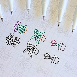 May include: A collection of white pens with black tips arranged around several small, colorful plant illustrations. The drawings include green, pink, and orange potted plants on a grid-patterned paper, suggesting a theme of art or stationery.