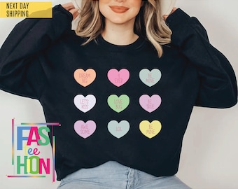 May include: A black long-sleeve sweatshirt featuring a grid of nine pastel-colored heart-shaped candies. Each heart contains text including DREAM BIG, 4 EVER, BE MINE, LETS KISS, LOVE BUG, BE TRUE, AWE SOME, LOL, and BE MINE in a simple sans-serif font.