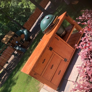 May include: A wooden outdoor kitchen island with a Big Green Egg grill. The island has a stained finish, cabinet doors, drawers, and a lower shelf. The grill is green with a silver top and sits on the island's surface.