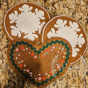 May include: Three brown felt Christmas decorations. Two are round with white glitter snowflake designs and white trim. The third is heart-shaped with green trim and candy embellishments. The decorations are festive and handmade.