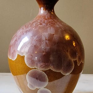 Może przedstawiać: Brązowy ceramiczny wazon o szerokim, zaokrąglonym korpusie i wąskiej szyjce. Wazon ma białą i fioletową glazurę z wirującym wzorem.