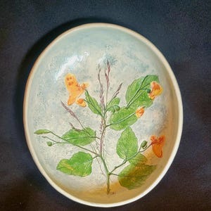 May include: A round ceramic bowl with a light blue and white mottled background. The center features a detailed botanical illustration of a plant with green leaves, thin stems, and small orange flowers with distinct petal shapes.