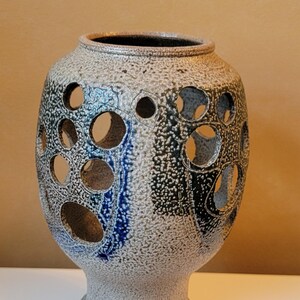 May include: A textured ceramic vase with a pedestal base and a wide, open top. The vase features large, circular cutouts and is primarily gray with blue and black accents. The vase is approximately 12 inches tall.