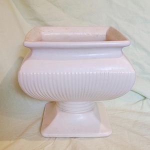 May include: A pale pink ceramic planter with a square opening and a flared rim. The body features vertical ribbed detailing around the center, supported by a square pedestal base. The surface has a smooth, glossy finish with a uniform light pink color.