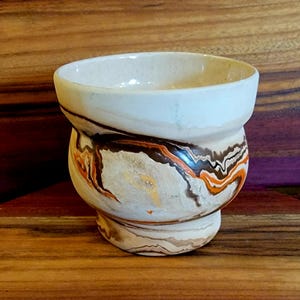 May include: A handmade ceramic bowl with a unique design. The bowl features a cream-colored rim and a body with swirling patterns of brown, orange, and cream. The bowl is set on a wooden surface.