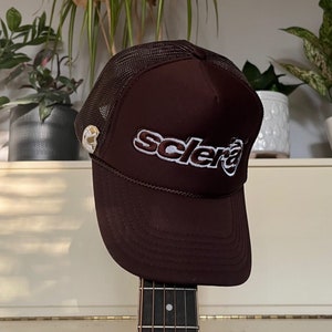 Könnte beinhalten: Eine braune Mesh-Trucker-Kappe mit einem weißen gestickten Logo, das vorne "scler" sagt. Die Kappe sitzt auf einem Gitarrenhals.