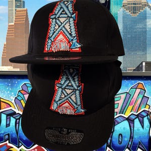 May include: Two black baseball caps, each with a colourful embroidered design. The design features a stylized letter "A" with blue, white, and red accents. The caps have a flat peak and a Cambridge label.