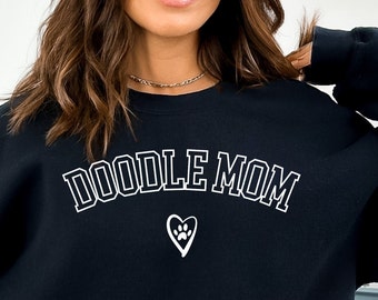 May include: Black sweatshirt with white text that reads "Doodle Mom" in a collegiate font. A white paw print inside a heart is below the text.