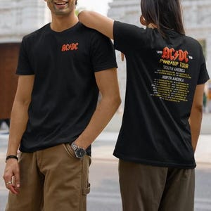 May include: A black short-sleeve t-shirt featuring the red AC/DC logo on the left chest. The back of the shirt displays the AC/DC PWR UP TOUR logo in red and yellow, followed by a list of tour dates and locations across South and North America.
