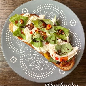 May include: A close-up of a toast topped with whipped ricotta cheese, fresh basil leaves, pine nuts, and a drizzle of balsamic glaze. The toast is on a gray plate with a white polka dot pattern. The text "@lajefasalsa" is at the bottom of the image.