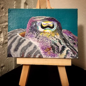 May include: A small painting on canvas of a purple and gray octopus with a yellow and black eye. The octopus is painted on a teal background and is displayed on a small wooden easel.