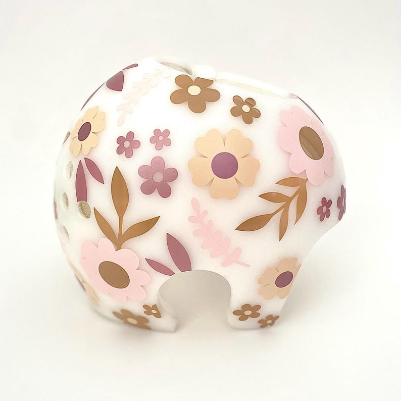May include: A white, floral-decorated piggy bank. The bank features a rounded shape with a slot on top and a curved opening at the bottom. The design includes pink, brown, and beige flowers and leaves.