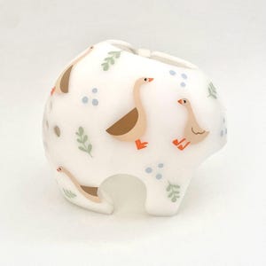 May include: White baby helmet decorated with cartoon geese in shades of brown, peach, and orange. The helmet also features green leaf and blue dot accents. The helmet has a rounded shape with a chin strap opening.