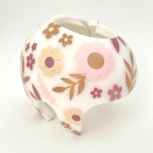 May include: White helmet decorated with pink, brown, and tan floral designs. The helmet has a rounded shape with a curved brim and a strap. The flowers are in various sizes and styles, with leaves and other floral accents.