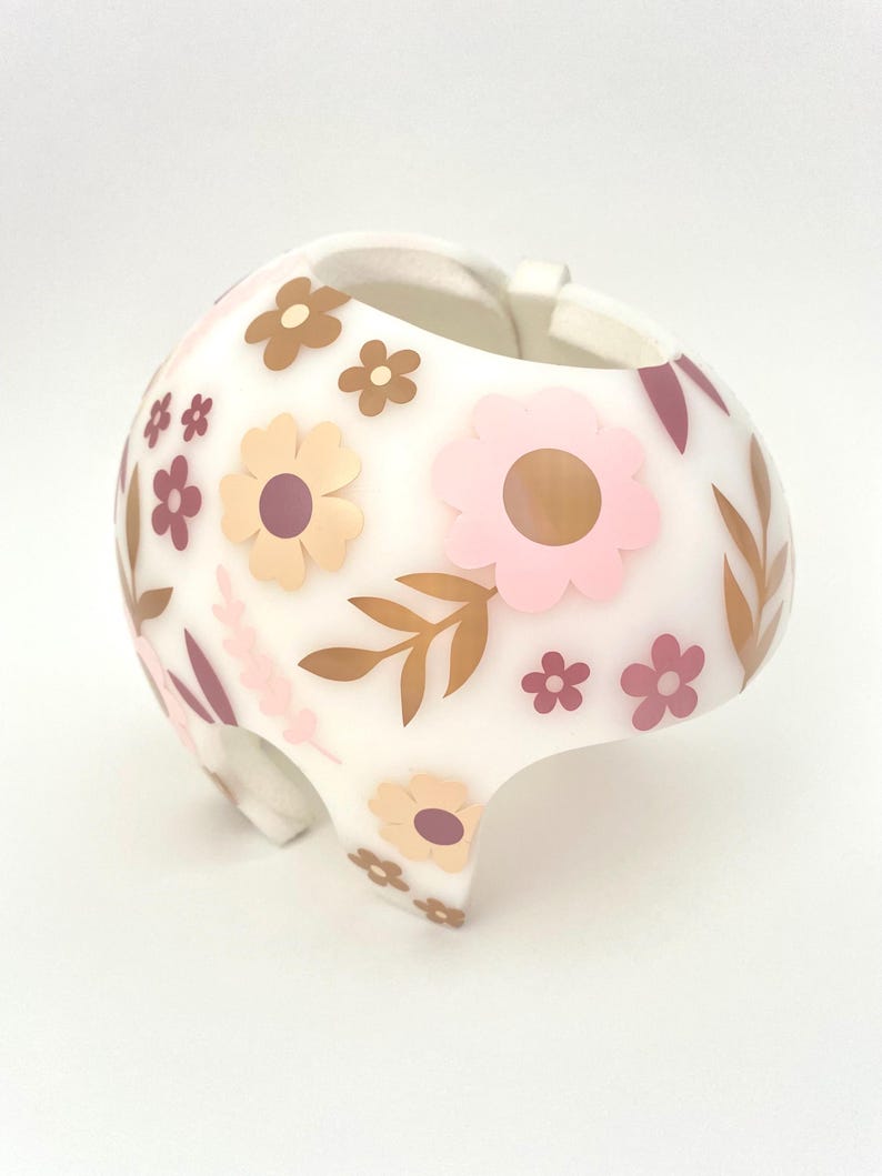 May include: White helmet decorated with pink, brown, and tan floral designs. The helmet has a rounded shape with a curved brim and a strap. The flowers are in various sizes and styles, with leaves and other floral accents.