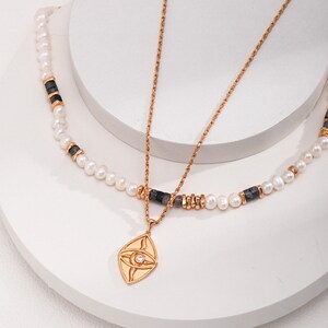 Könnte beinhalten: Eine goldfarbene Halskette mit einem Anhänger in Augenform und einem kleinen, klaren Stein. Die Halskette ist mit einer Perlen- und Stein-Halskette kombiniert. Der Schmuck wird auf einer weißen Oberfläche präsentiert.