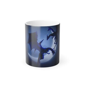 May include: A white ceramic mug with a wraparound design featuring dark blue silhouettes of dragons against a blue background with a crescent moon. The mug has a glossy finish and a white interior.