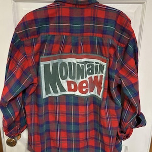 May include: Red, green, and blue plaid flannel shirt with a Mountain Dew logo patch on the back. The long-sleeved shirt has a button-down collar and cuffs. The Mountain Dew logo is in white and green.