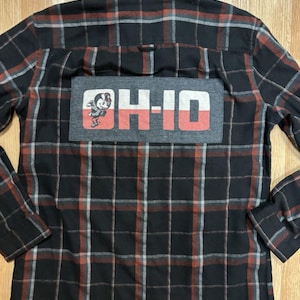 May include: A long-sleeve button-down flannel shirt in a black, red, and grey plaid pattern. The back features a rectangular grey patch with the text OH-IO in white and red, accompanied by a small graphic of a cartoon character wearing a hat.