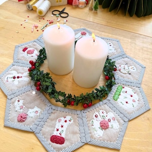 May include: Two white pillar candles with lit flames sit on a Christmas-themed centrepiece. The centrepiece features a green garland with red berries, surrounded by a quilted, hexagonal mat with festive embroidered designs and a striped border.