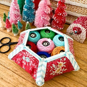 Peut inclure: Une trousse de couture en tissu rouge, blanc et bleu remplie de bobines de fil colorées. La trousse est entourée de sapins de Noël miniatures en forme de goupillon de différentes couleurs et d'une paire de ciseaux noirs.