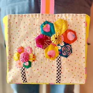 Könnte beinhalten: Eine handgefertigte Tragetasche mit floralem Design. Die Tasche hat einen cremefarbenen Hintergrund mit bunten Sternmustern und zwei großen Blumenapplikationen. Die Blumen bestehen aus verschiedenen gemusterten Stoffen, darunter Erdbeeren und Rosen. Die Tasche hat einen gelben Rand und einen rosa-orangenen Riemen.