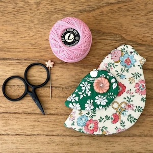 May include: A pink ball of Perle Arco Iris thread, a pair of black embroidery scissors, a star-topped sewing pin, and a bird-shaped fabric pincushion with a floral pattern, a green wing, and a small pink button on the wing.