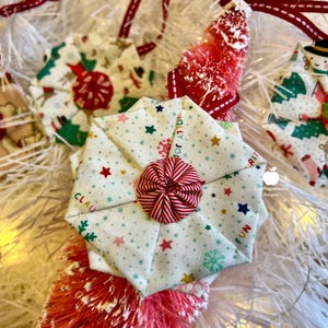 Könnte beinhalten: Ein weißer Stoff-Weihnachtsbaumschmuck mit einem rot-weiß gestreiften Knopf in der Mitte. Der Schmuck ist mit einem weißen Hintergrund mit kleinen blauen Schneeflocken, roten Sternen und goldenen Sternen verziert. Der Stoff hat die Wörter "Jolly", "Claus" und "Fun" aufgedruckt.