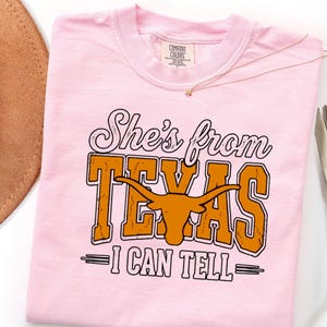 Pu&ograve; includere: Una t-shirt rosa chiaro con il testo 'She's from TEXAS I CAN TELL' in un mix di caratteri corsivi e stampatello. La parola 'TEXAS' &egrave; arancione con una texture usurata e include un grafico di una testa di longhorn al centro.