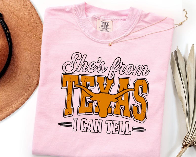 Pu&ograve; includere: Una t-shirt rosa chiaro con il testo 'She's from TEXAS I CAN TELL' in un mix di caratteri corsivi e stampatello. La parola 'TEXAS' &egrave; arancione con una texture usurata e include un grafico di una testa di longhorn al centro.