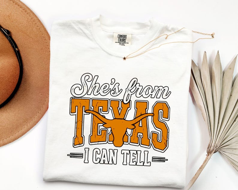 Pu&ograve; includere: Una t-shirt bianca a maniche corte con una stampa grafica che recita She's from TEXAS I CAN TELL in caratteri neri e arancioni, con una silhouette centrale di un toro longhorn arancione. La maglietta presenta un'etichetta Comfort Colors.