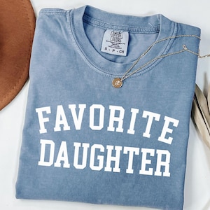 Peut inclure: Un t-shirt bleu Comfort Colors avec les mots "FAVORITE DAUGHTER" imprim&eacute;s en lettres blanches. Un collier en or avec un pendentif repose sur le t-shirt. Un chapeau marron et une plante s&eacute;ch&eacute;e sont en arri&egrave;re-plan.