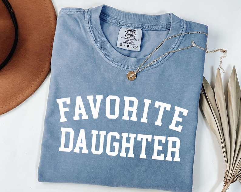 Peut inclure: Un t-shirt bleu Comfort Colors avec les mots "FAVORITE DAUGHTER" imprim&eacute;s en lettres blanches. Un collier en or avec un pendentif repose sur le t-shirt. Un chapeau marron et une plante s&eacute;ch&eacute;e sont en arri&egrave;re-plan.