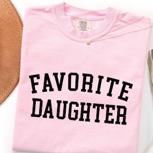 Peut inclure: T-shirt rose clair avec l'inscription "FAVORITE DAUGHTER" en lettres noires grasses. Un collier dor&eacute; et un chapeau marron sont visibles en arri&egrave;re-plan. Le t-shirt porte une &eacute;tiquette Comfort Colors.