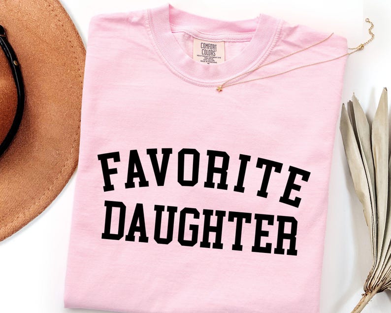 Peut inclure: T-shirt rose clair avec l'inscription "FAVORITE DAUGHTER" en lettres noires grasses. Un collier dor&eacute; et un chapeau marron sont visibles en arri&egrave;re-plan. Le t-shirt porte une &eacute;tiquette Comfort Colors.