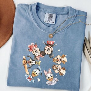 May include: A blue Comfort Colors t-shirt with a circular watercolour design of Disney characters, including Mickey and Minnie Mouse, Goofy, Donald and Daisy Duck, and Chip and Dale. A gold necklace rests on the shirt.