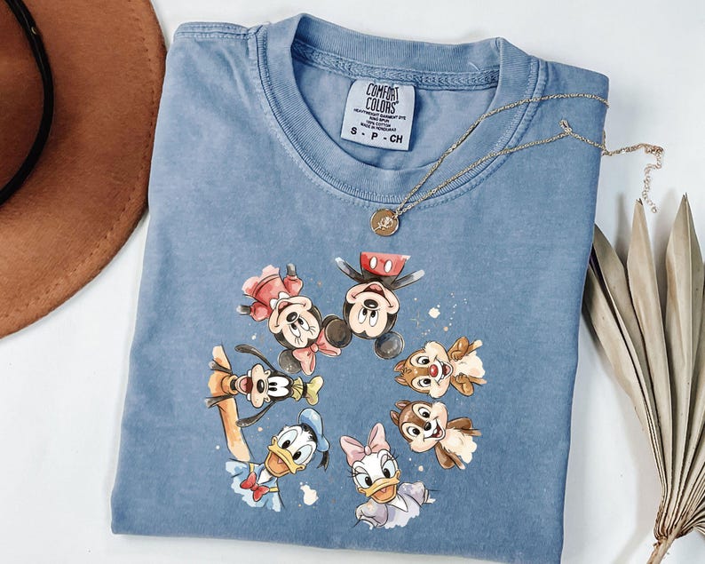 May include: A blue Comfort Colors t-shirt with a circular watercolour design of Disney characters, including Mickey and Minnie Mouse, Goofy, Donald and Daisy Duck, and Chip and Dale. A gold necklace rests on the shirt.