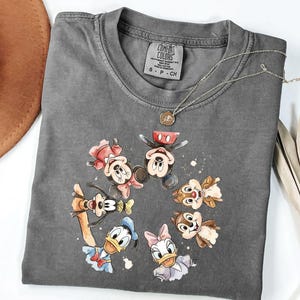 May include: A grey Comfort Colors t-shirt with a watercolour design of Mickey Mouse, Minnie Mouse, Donald Duck, Daisy Duck, Goofy, and Chip and Dale. A silver necklace with a small pendant rests on the shirt.