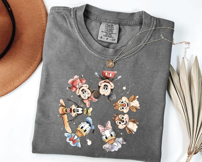 May include: A grey Comfort Colors t-shirt with a watercolour design of Mickey Mouse, Minnie Mouse, Donald Duck, Daisy Duck, Goofy, and Chip and Dale. A silver necklace with a small pendant rests on the shirt.