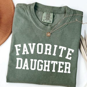 Peut inclure: Un t-shirt vert olive avec les mots "FAVORITE DAUGHTER" imprim&eacute;s en lettres blanches. Un collier en or avec un pendentif repose sur le t-shirt. Le t-shirt est pli&eacute; sur une surface blanche.