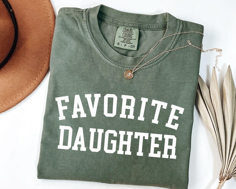 Peut inclure: Un t-shirt vert olive avec les mots "FAVORITE DAUGHTER" imprim&eacute;s en lettres blanches. Un collier en or avec un pendentif repose sur le t-shirt. Le t-shirt est pli&eacute; sur une surface blanche.