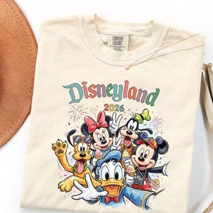 Może przedstawiać: Kremowy t-shirt z kolorową grafiką przedstawiającą Myszkę Miki, Myszkę Minnie, Kaczora Donalda, Goofy'ego i Pluto. Napis głosi Disneyland 2026. Postacie są przedstawione z rożkami lod&oacute;w i aparatem fotograficznym, w otoczeniu fajerwerk&oacute;w.