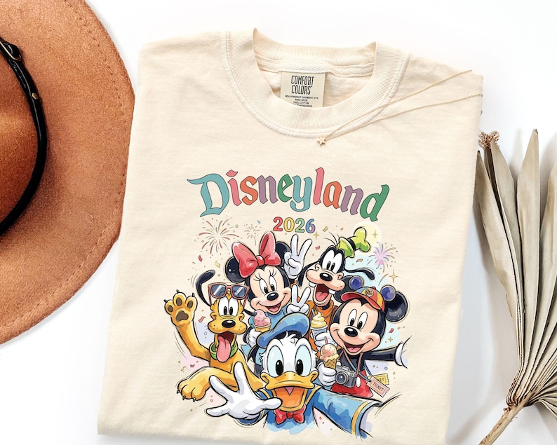 Może przedstawiać: Kremowy t-shirt z kolorową grafiką przedstawiającą Myszkę Miki, Myszkę Minnie, Kaczora Donalda, Goofy'ego i Pluto. Napis głosi Disneyland 2026. Postacie są przedstawione z rożkami lod&oacute;w i aparatem fotograficznym, w otoczeniu fajerwerk&oacute;w.
