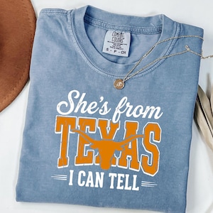 Pu&ograve; includere: Una t-shirt blu a maniche corte con una stampa grafica bianca e arancione che recita She's from TEXAS I CAN TELL, con una silhouette di un bue longhorn arancione integrata nella parola TEXAS. La maglietta presenta un girocollo e taglia S.