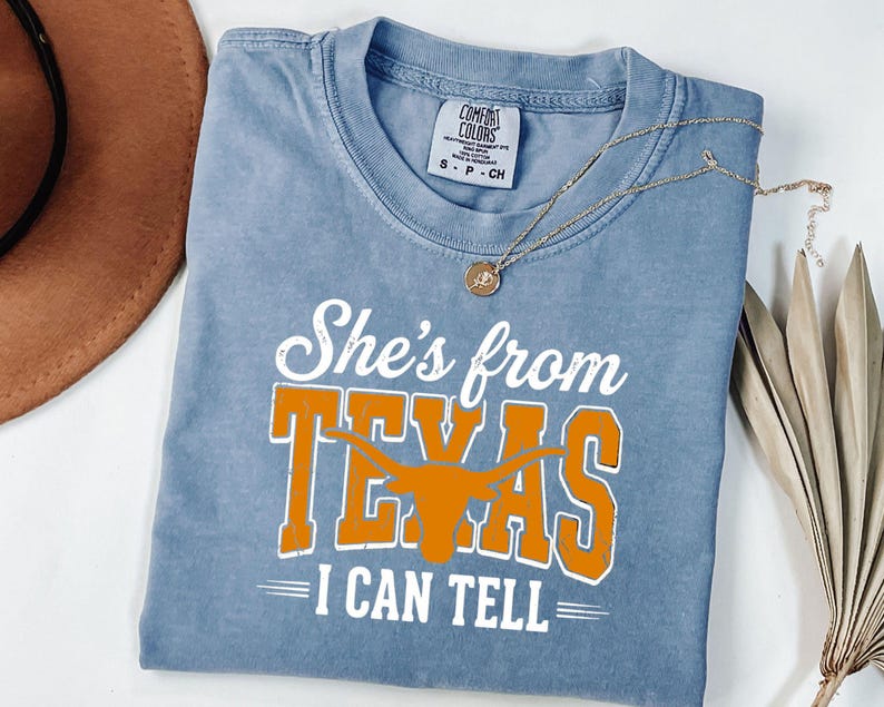 Pu&ograve; includere: Una t-shirt blu a maniche corte con una stampa grafica bianca e arancione che recita She's from TEXAS I CAN TELL, con una silhouette di un bue longhorn arancione integrata nella parola TEXAS. La maglietta presenta un girocollo e taglia S.