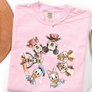 May include: A pale pink Comfort Colors sweatshirt with a watercolour design of Mickey Mouse, Minnie Mouse, Donald Duck, Daisy Duck, Goofy, and Chip and Dale. The characters are arranged in a circle. A gold necklace and a brown hat are in the background.