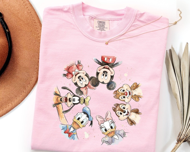 May include: A pale pink Comfort Colors sweatshirt with a watercolour design of Mickey Mouse, Minnie Mouse, Donald Duck, Daisy Duck, Goofy, and Chip and Dale. The characters are arranged in a circle. A gold necklace and a brown hat are in the background.