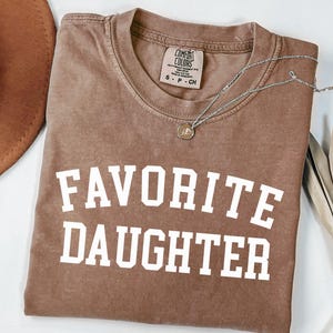 Peut inclure: Un t-shirt marron Comfort Colors avec les mots "FAVORITE DAUGHTER" imprim&eacute;s en lettres blanches. Un collier en argent avec un pendentif est drap&eacute; sur le t-shirt. Un chapeau marron et une plante s&eacute;ch&eacute;e sont en arri&egrave;re-plan.