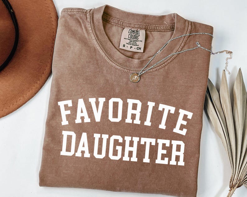 Peut inclure: Un t-shirt marron Comfort Colors avec les mots "FAVORITE DAUGHTER" imprim&eacute;s en lettres blanches. Un collier en argent avec un pendentif est drap&eacute; sur le t-shirt. Un chapeau marron et une plante s&eacute;ch&eacute;e sont en arri&egrave;re-plan.
